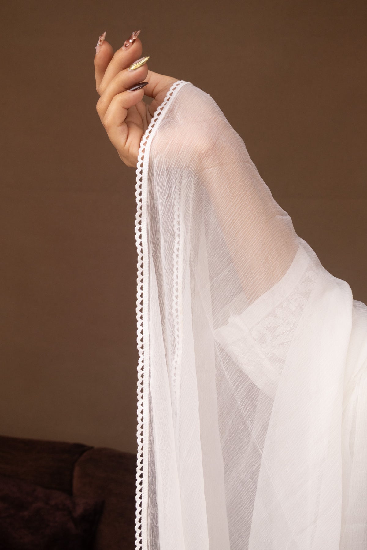 A close-up of a person's hand holding up a sheer white fabric sleeve or drape with a delicate scalloped edge, set against a warm brown background.