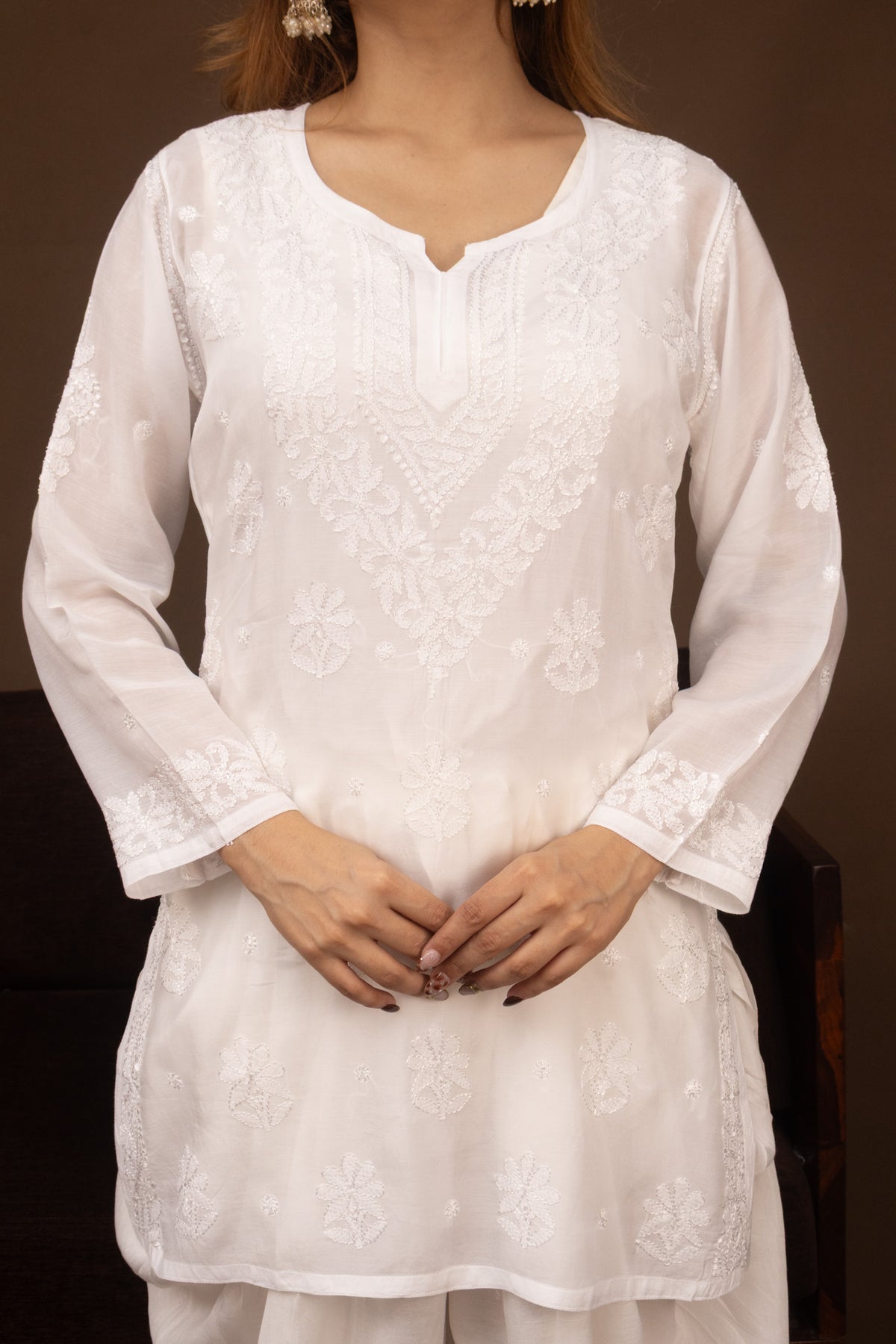 A woman wearing a white embroidered kurta with long sleeves, a split neckline, and delicate floral embroidery, paired with matching white trousers against a dark brown background.