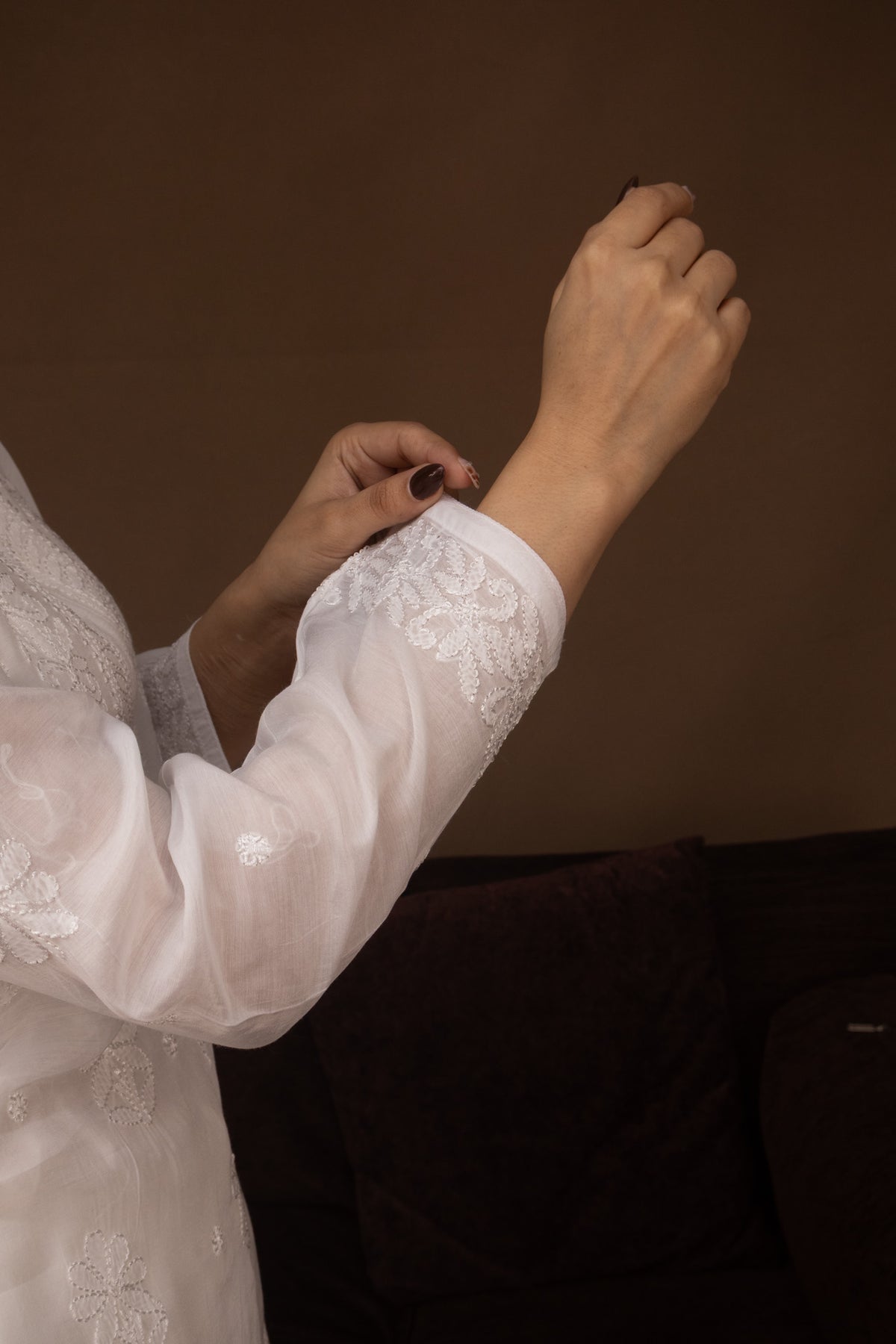 A close-up of a person wearing a white embroidered traditional garment, adjusting the sleeve with one hand while the other hand rests near the wrist. The background is a dark brown wall and a dark sofa.
