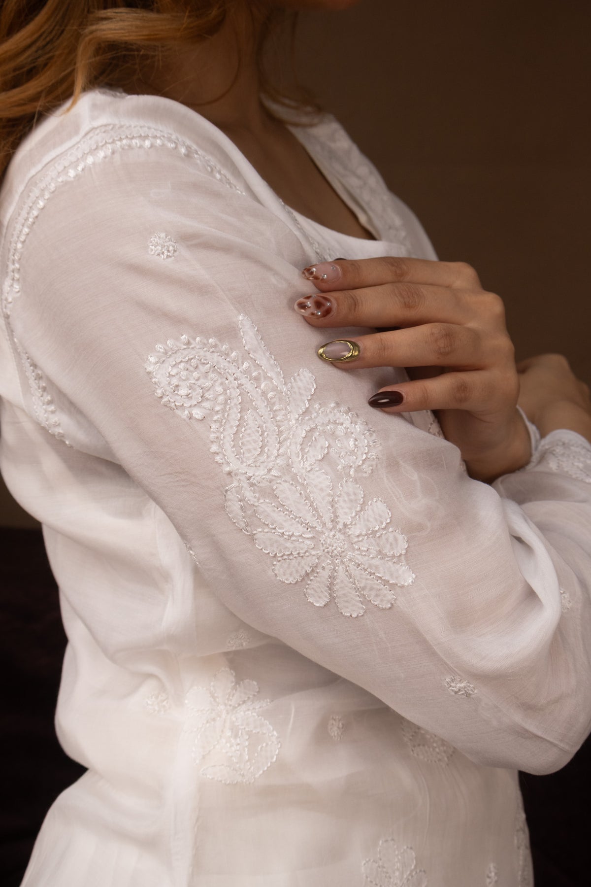 A close-up of a woman wearing a white semi-sheer embroidered robe or kimono-style top with floral lace detailing on the sleeve, against a dark brown background.