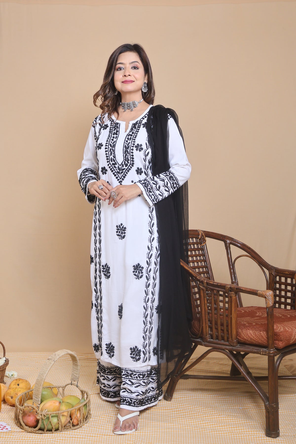 A woman models a white traditional South Asian outfit with black floral embroidery, a black dupatta, and matching black-and-white trousers, standing in front of a beige studio backdrop next to a brown woven chair.