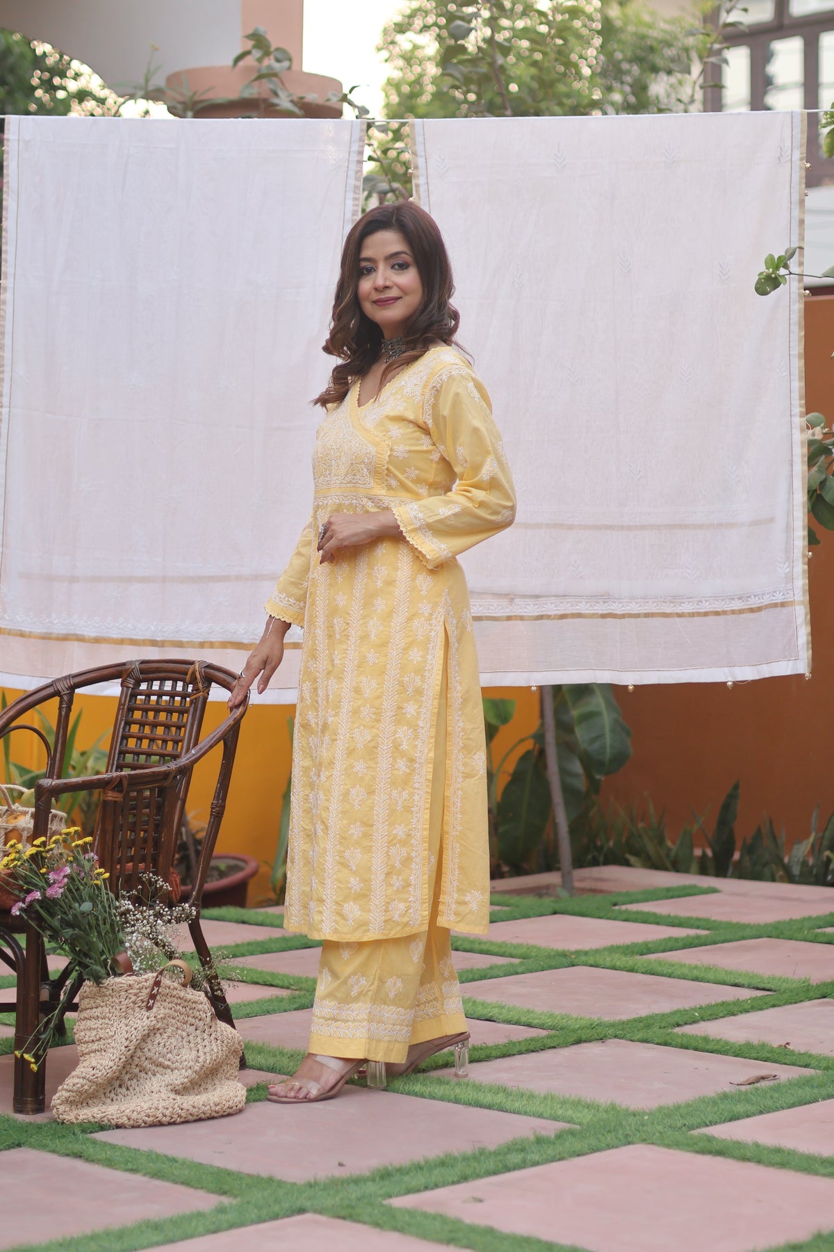 A woman wearing a yellow embroidered kurta and matching pants stands in an outdoor courtyard with a white hanging cloth behind her.