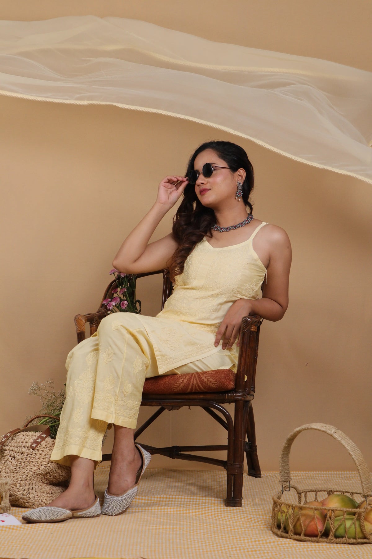 A woman wearing a light yellow sleeveless kurta and matching trousers sits on a wooden chair in a studio setting with a beige backdrop, draped sheer fabric overhead, and baskets on the floor.