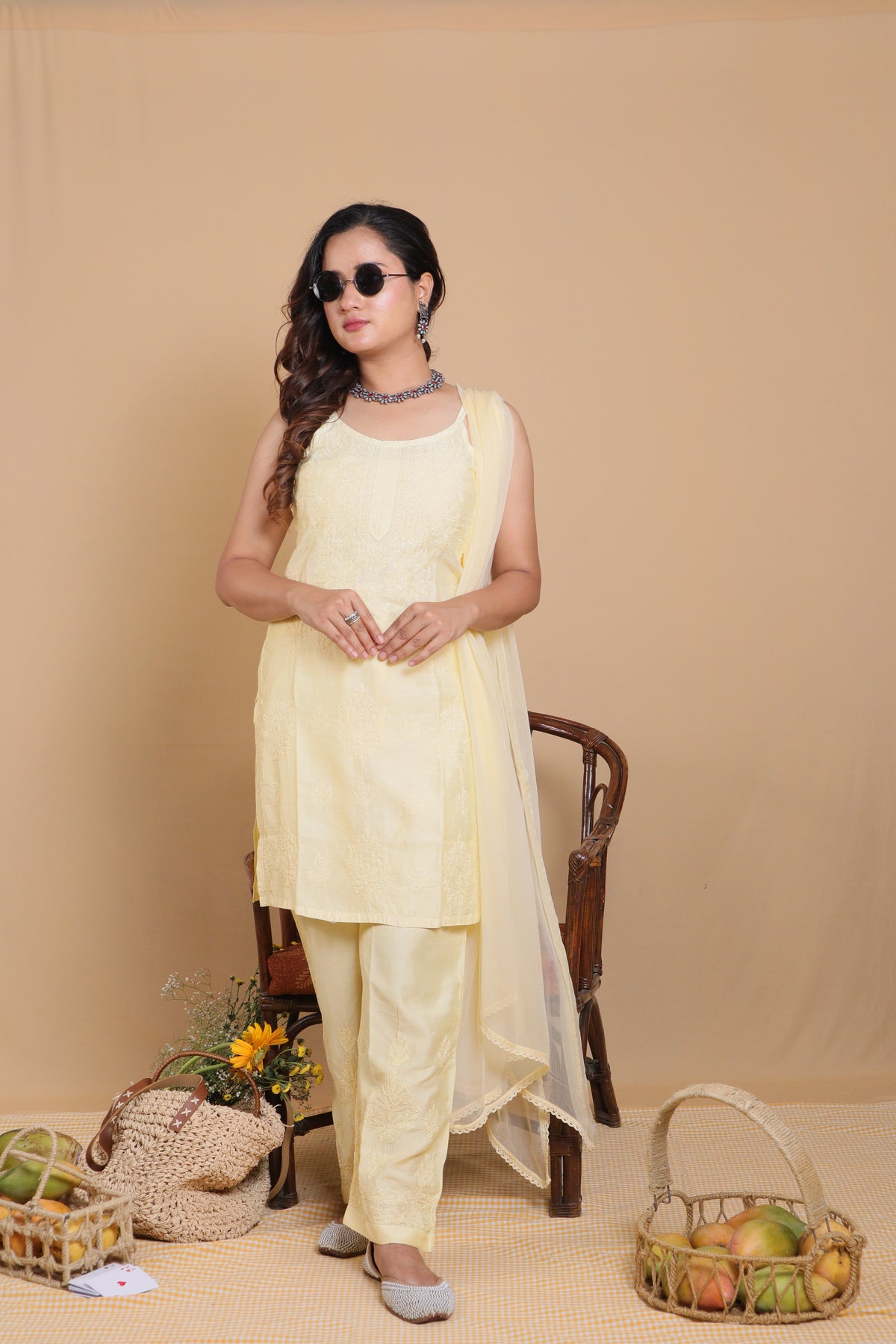 A woman modeling a light yellow traditional Indian salwar kameez set with a matching dupatta, standing in front of a beige backdrop with decorative baskets and a chair.