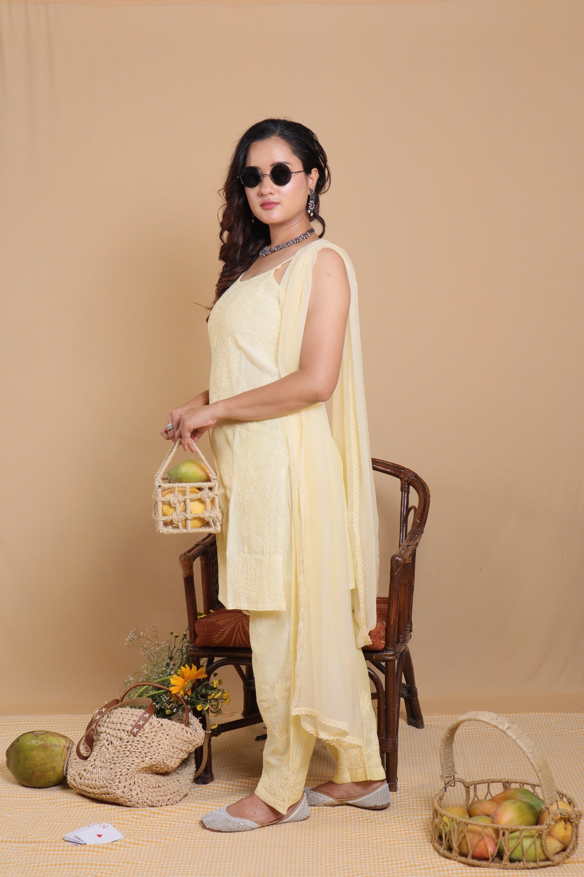 A woman modeling a light yellow traditional Indian salwar kameez suit, standing next to a wooden chair in a studio setting with decorative props like a woven bag, fruit baskets, and a coconut.