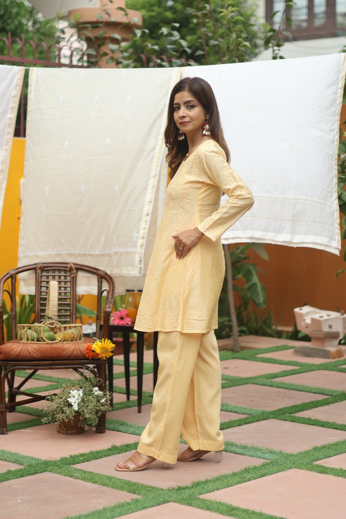 A woman modeling a light yellow kurta and trouser set in an outdoor patio setting with a white sheet hanging in the background.