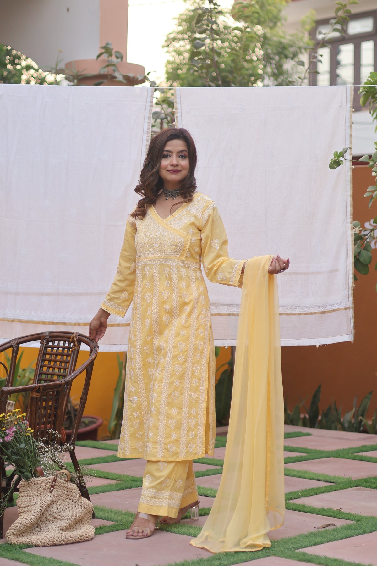 A woman models a light yellow ethnic salwar kameez suit with matching embroidered pants and a sheer dupatta, set against a background with hanging white sheets and a wicker chair.