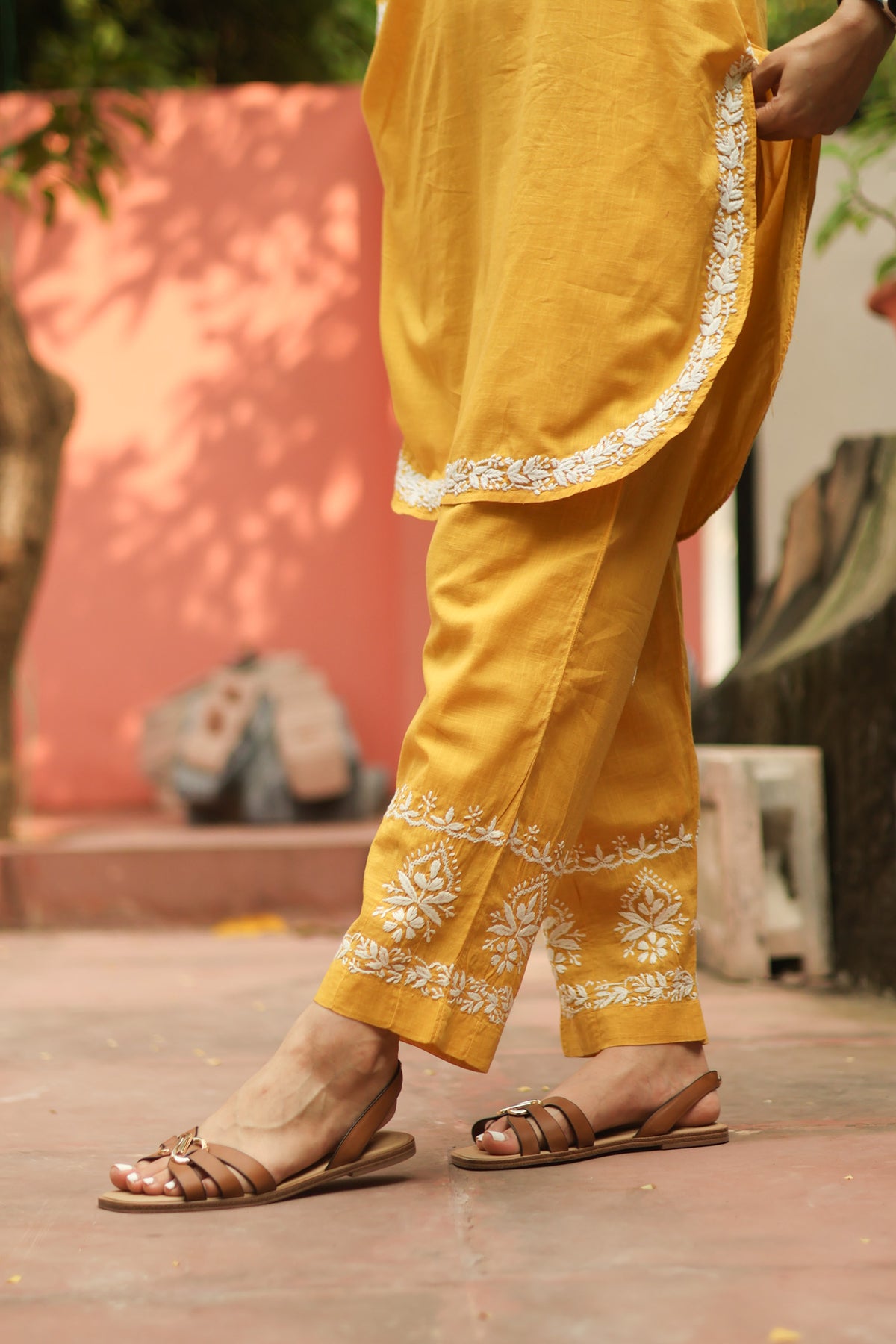 A person wearing a mustard yellow traditional outfit with white floral embroidery along the hem and kurta border, paired with brown leather slide sandals, standing outdoors against a peach-colored wall.