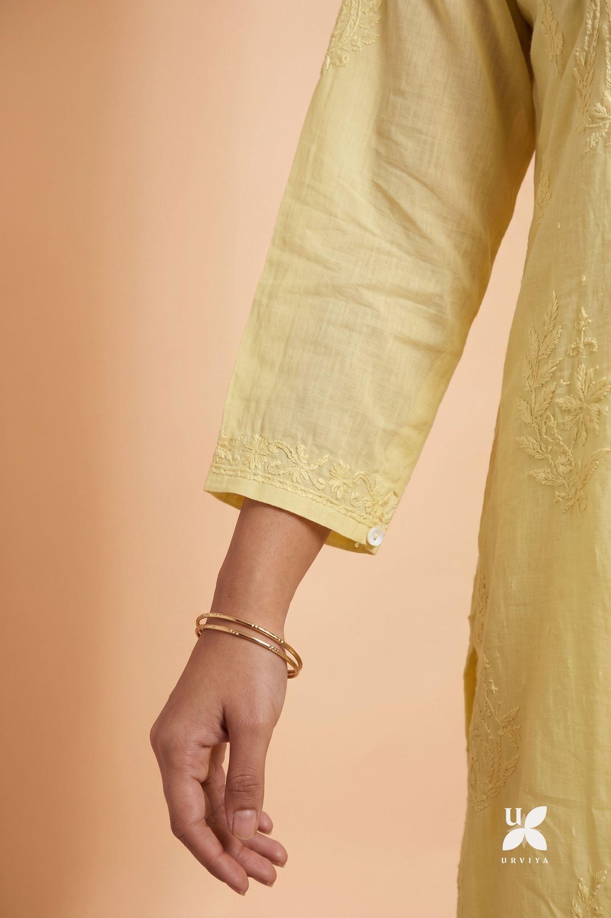 A close-up of a person's hand and wrist wearing a light yellow embroidered kurta or dress, with a gold bangle, against a solid peach background. The Uniqlo logo is visible in the bottom right corner.