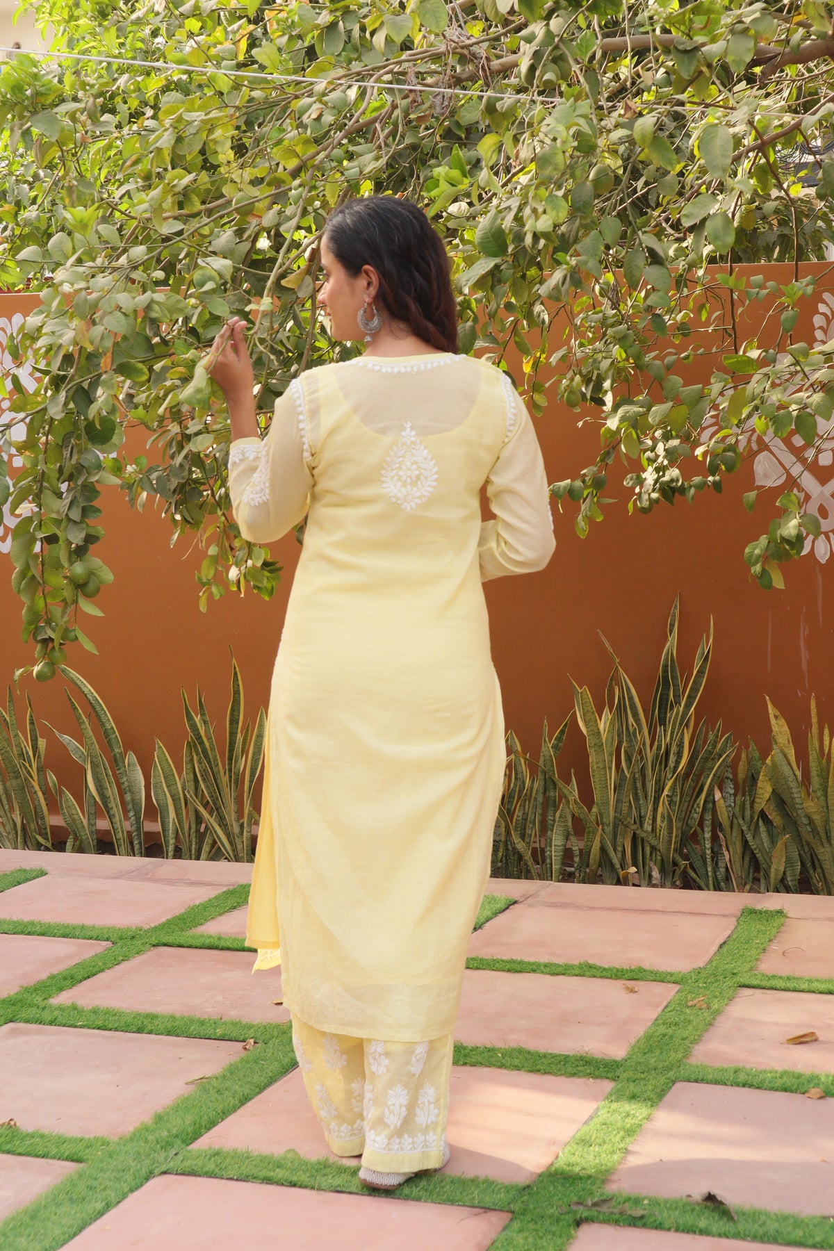 A woman wearing a light yellow ethnic suit with white embroidery, photographed from behind in an outdoor garden setting with a terracotta wall and green plants.