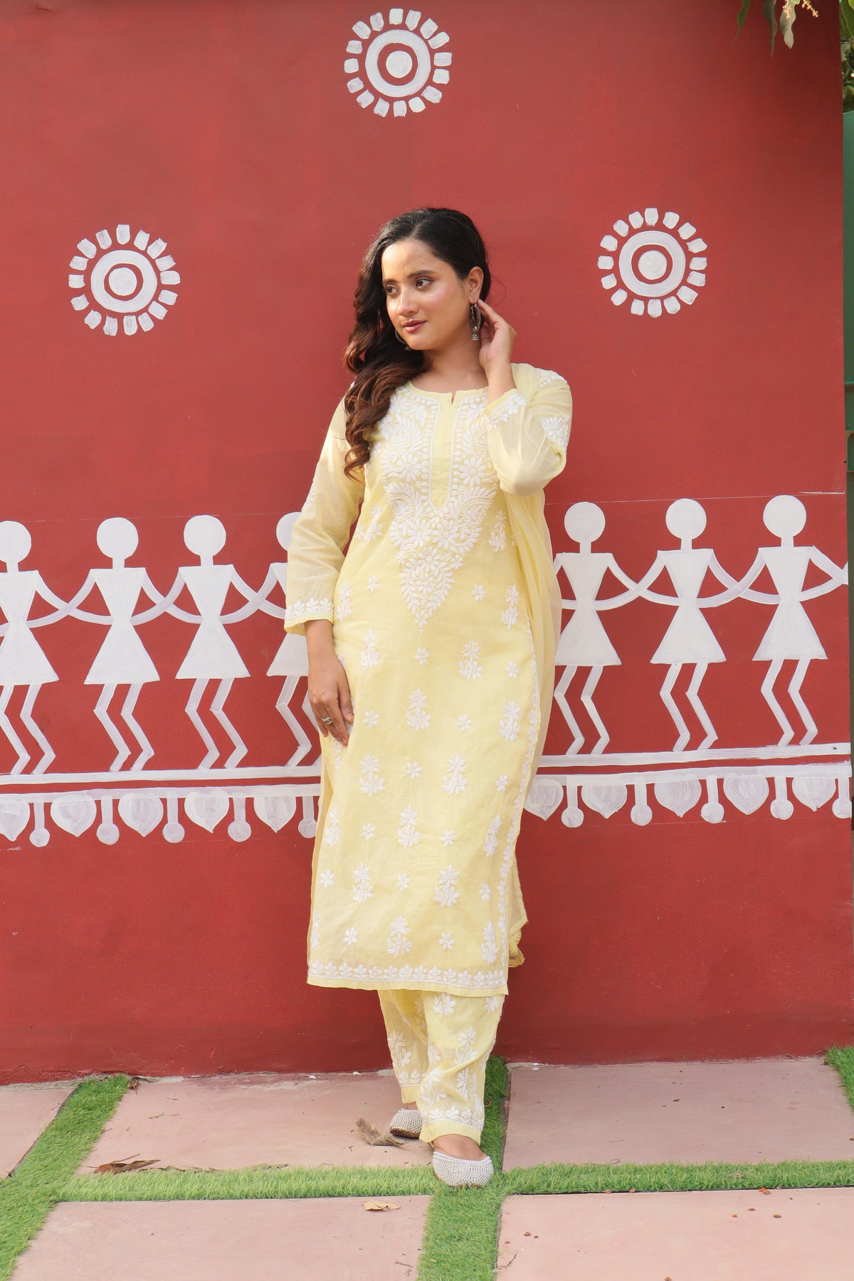 A woman modeling a light yellow traditional Indian salwar kameez suit with white floral embroidery, standing in front of a red wall decorated with white folk art.