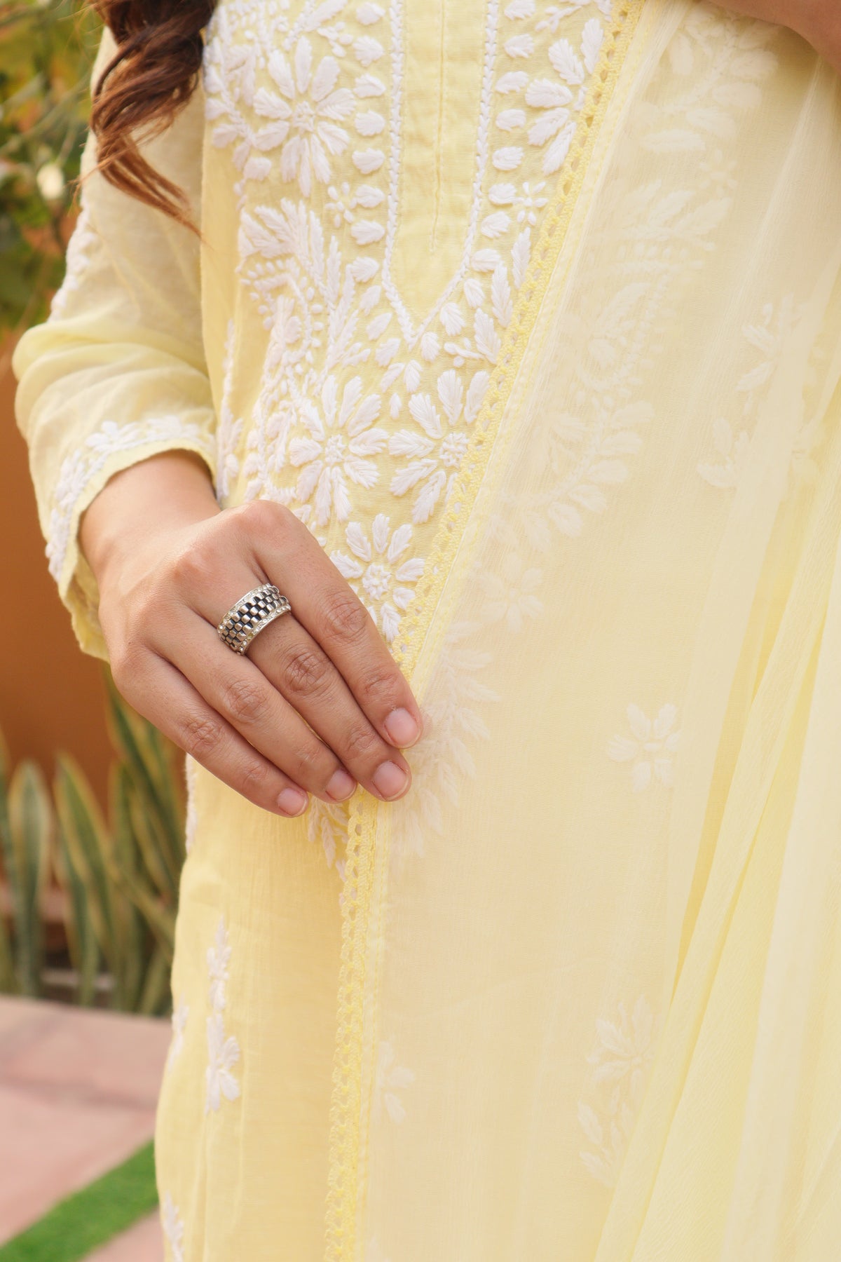A close-up of a person wearing a pale yellow embroidered kurta with white floral embroidery and a matching sheer dupatta, shown outdoors with a blurred background.