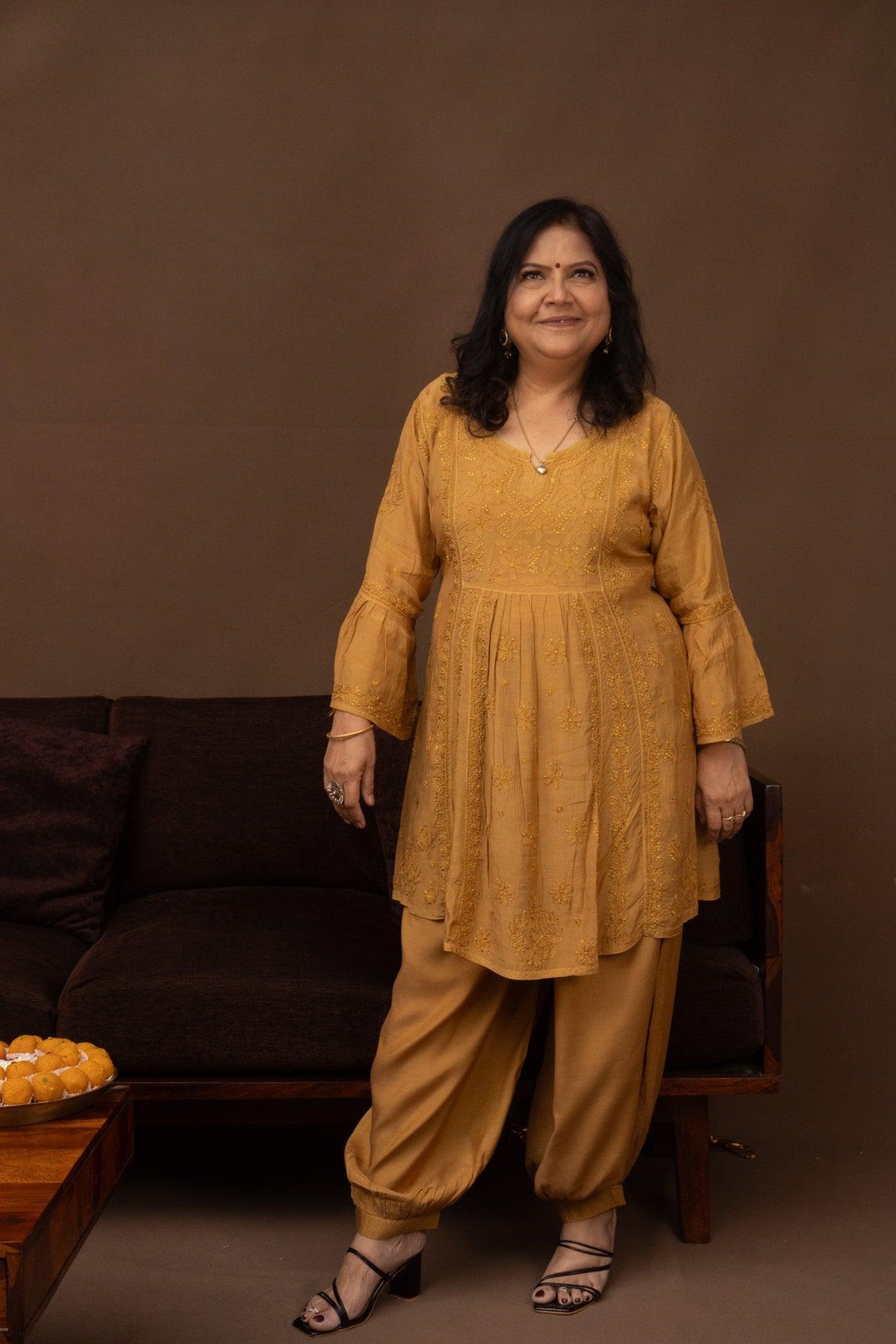 A woman wearing a mustard yellow embroidered kurta with matching wide-leg pants, standing in front of a brown sofa against a solid brown backdrop. Yellow Muslin Nyra Cut Chikankari Kurti with Bakhiya Embroidery