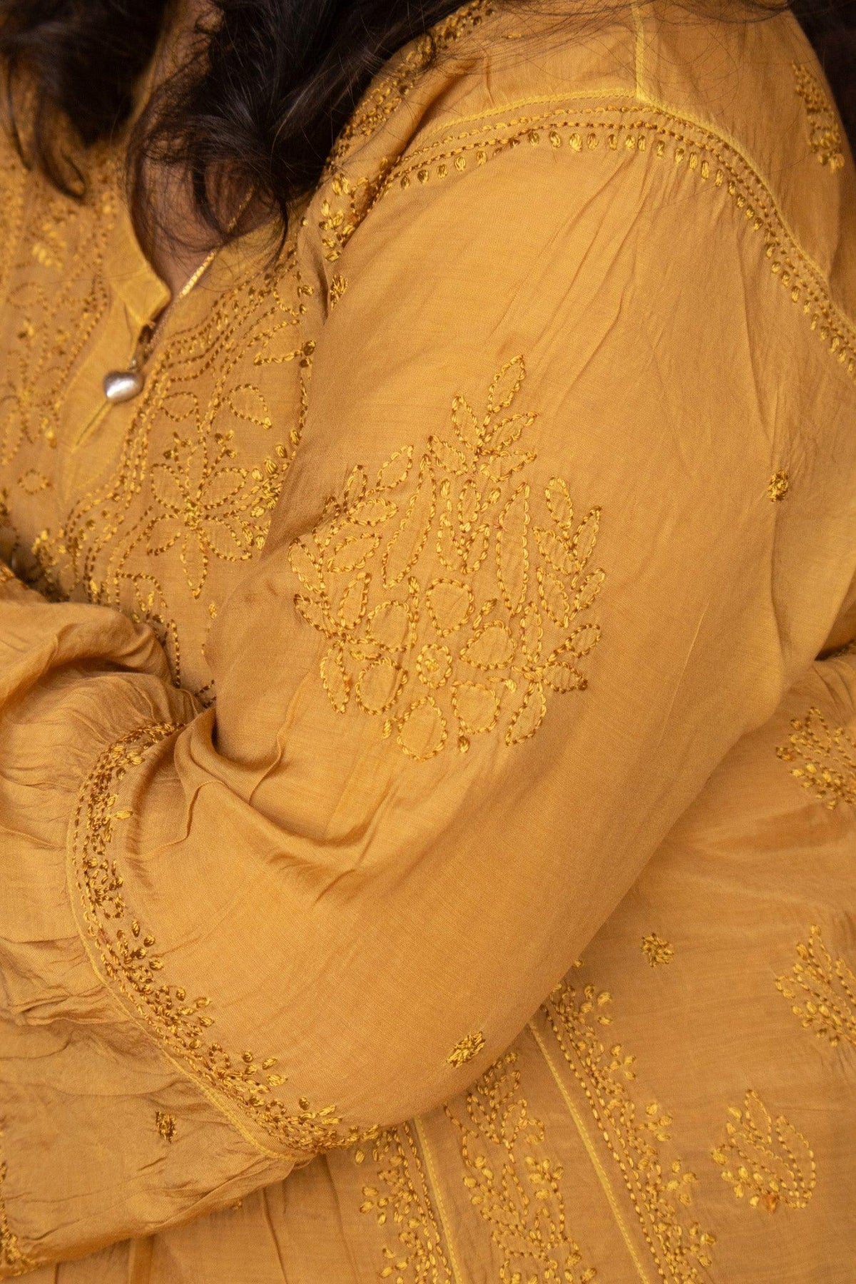 Close-up of a mustard yellow embroidered fabric garment, showing detailed floral embroidery on the sleeve and bodice with a pearl-like button. Yellow Muslin Nyra Cut Chikankari Kurti with Bakhiya Embroidery