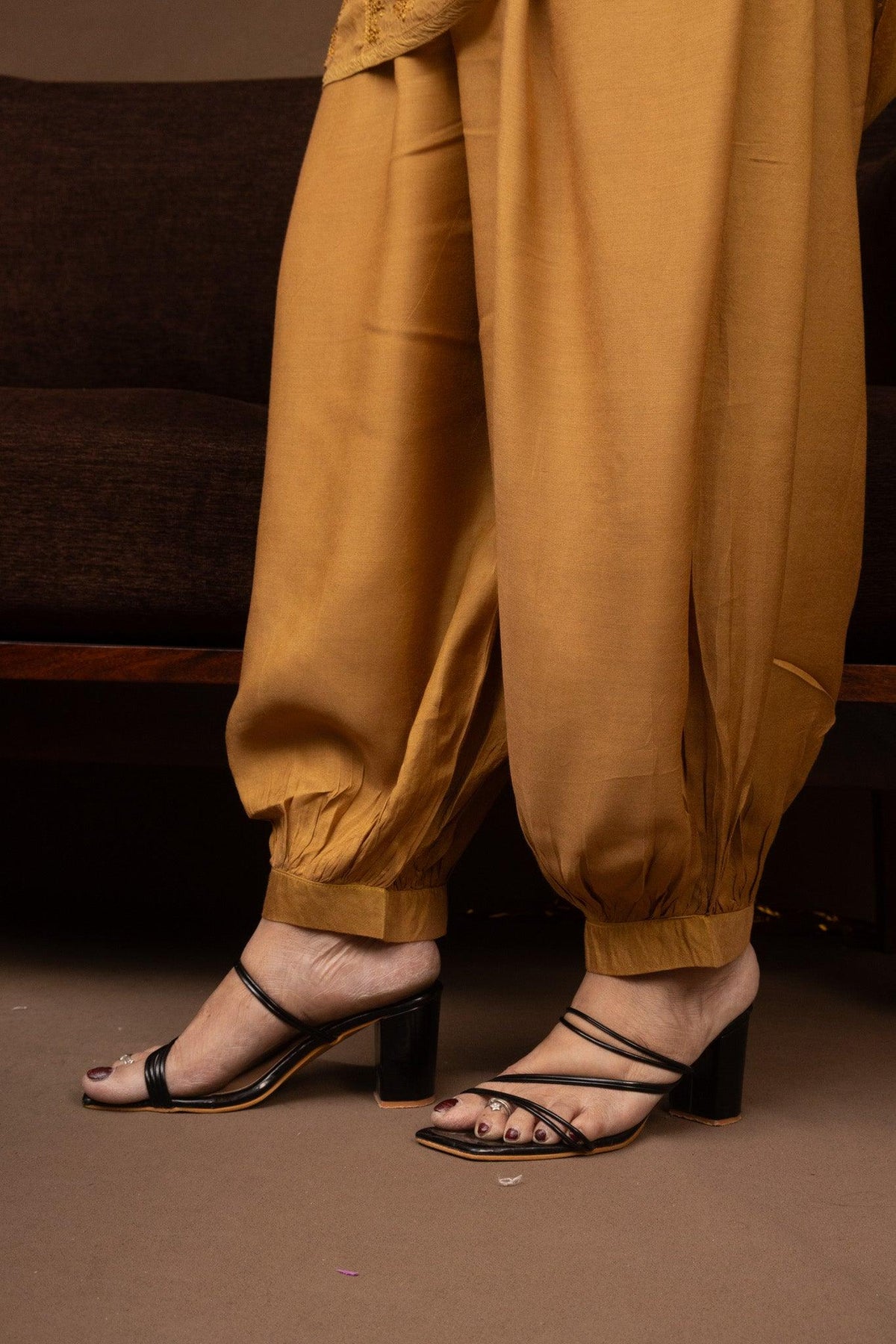 A close-up of a person's feet wearing mustard yellow harem pants and black strappy block-heel sandals, standing indoors on a brown floor. Yellow Muslin Nyra Cut Chikankari Kurti with Bakhiya Embroidery