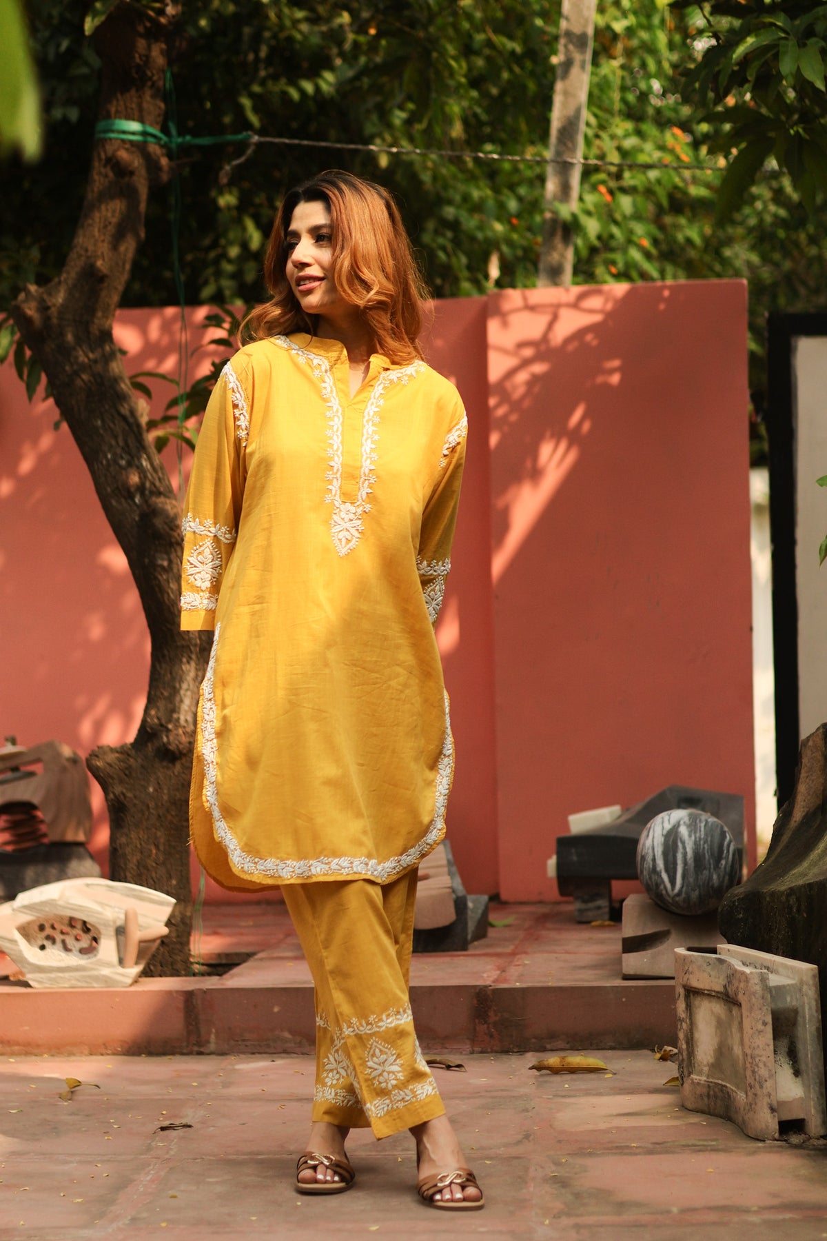 A woman wearing a mustard yellow traditional kurta and pant set with white embroidery, standing outdoors in front of a pink wall and green trees.