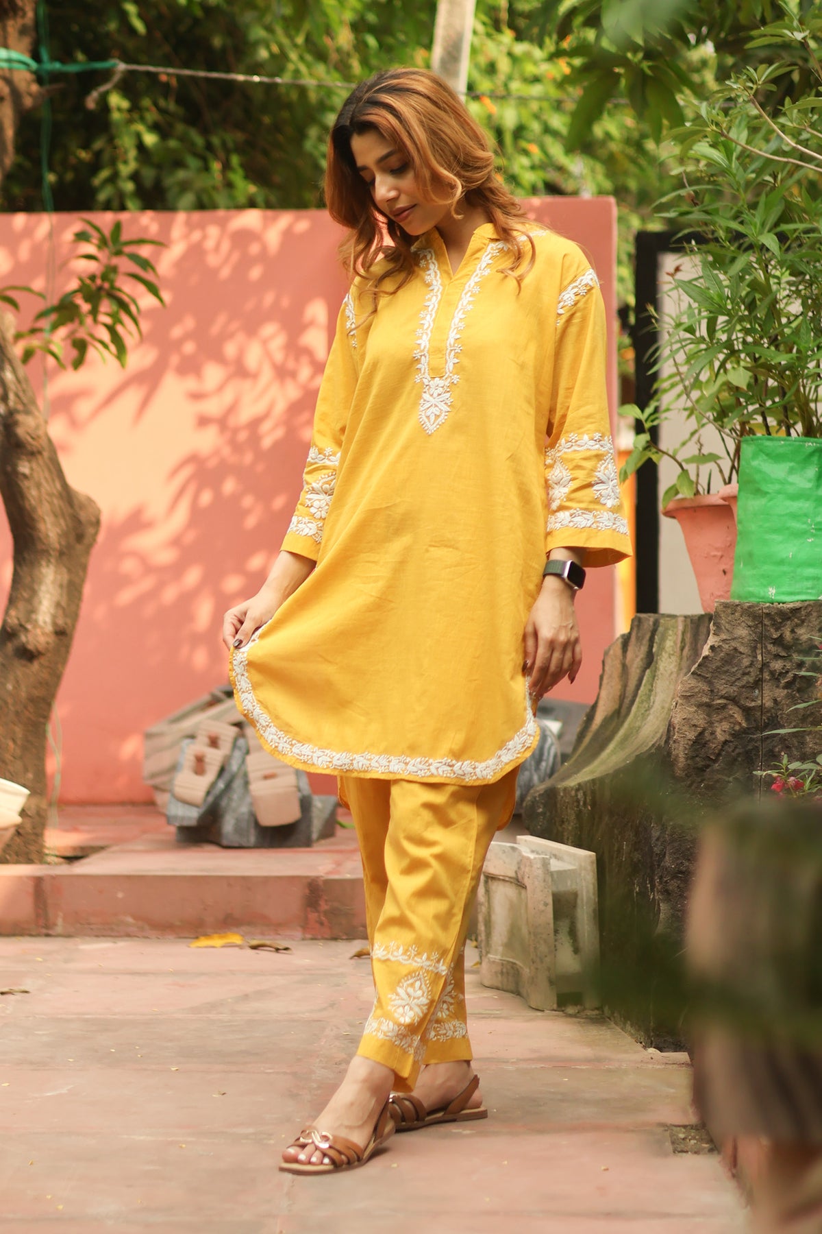 A woman wearing a bright yellow traditional kurta and matching pants with white embroidered details, standing outdoors on a paved path with a pink wall and green plants in the background.